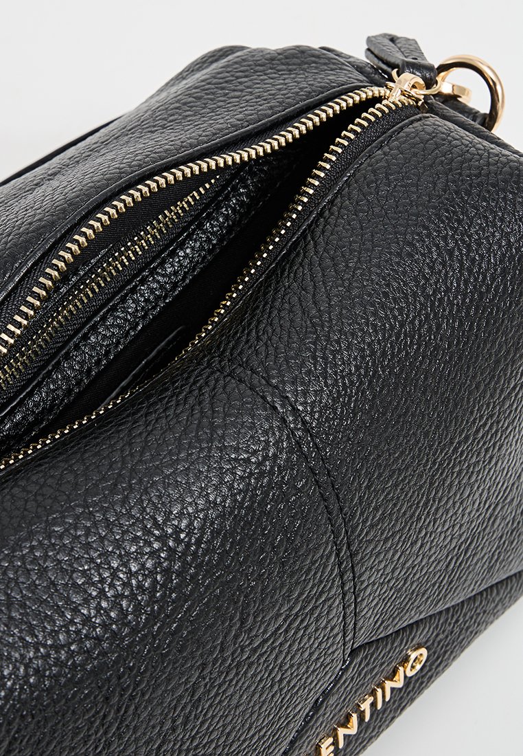 Sac à main en cuir noir texturé avec une fermeture éclair dorée partiellement ouverte et une inscription en doré sur fond blanc.