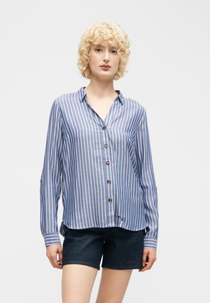 Nainen, jolla on kiharat vaaleat hiukset, pukeutunut sinivalkoraidalliseen nappipaitaan ja tummiin shortseihin yksinkertaisen vaalean taustan edessä.