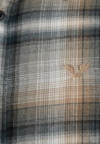 Camisa de cuadros en tonos azules, grises y beige. El material es una tela tejida con una textura suave. Presenta un logo de ala bordado en el pecho.