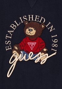 Sudadera de color navy con un oso que lleva una sudadera roja con un logo, texto bordado "Establecido en 1981" y "guess" en bordado ligero.