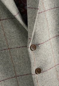 Szara marynarka z wełny w kratę nośną, z subtelnym wzorem w czerwone i brązowe paski. Posiada klapę z wycięciem oraz brązowe guziki. Tkanina o fakturze.