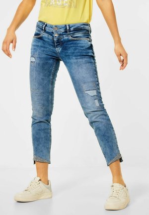 Person iført afklippede blå slidte jeans med dekorative nitter, gul skjorte og hvide sneakers, stående mod en ensfarvet baggrund.