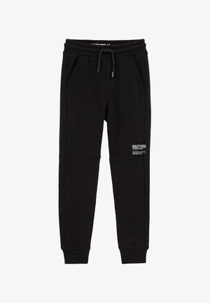 Juodos sportinės kelnės su elastinga liemene, raišteliu, šoninėmis kišenėmis, banguotais rankogaliais ir etikete dešinėje šlaunies pusėje.