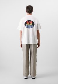 Hvit bomull t-skjorte med et fargerikt sirkulært grafikkdesign på ryggen. Modellen har på seg løse beige bukser og sportssko.