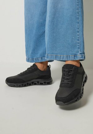 Person iført blå jeans med vide ben og sorte snørefede sneakers med luftpolstrede såler, der står på en lys gulv.