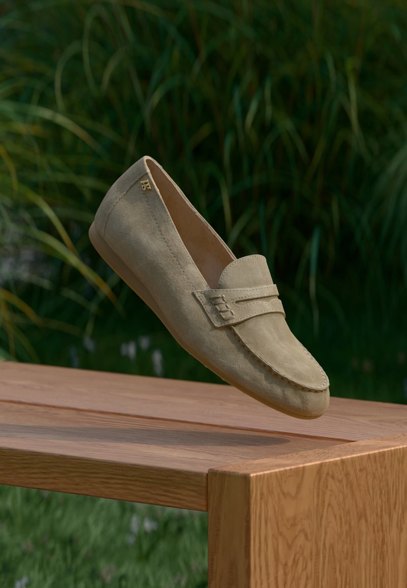 Mocassin beige en daim avec une semelle intérieure rembourrée, présentant un bout cousu et une bride décorative à l'avant, suspendu au-dessus d'une surface en bois.