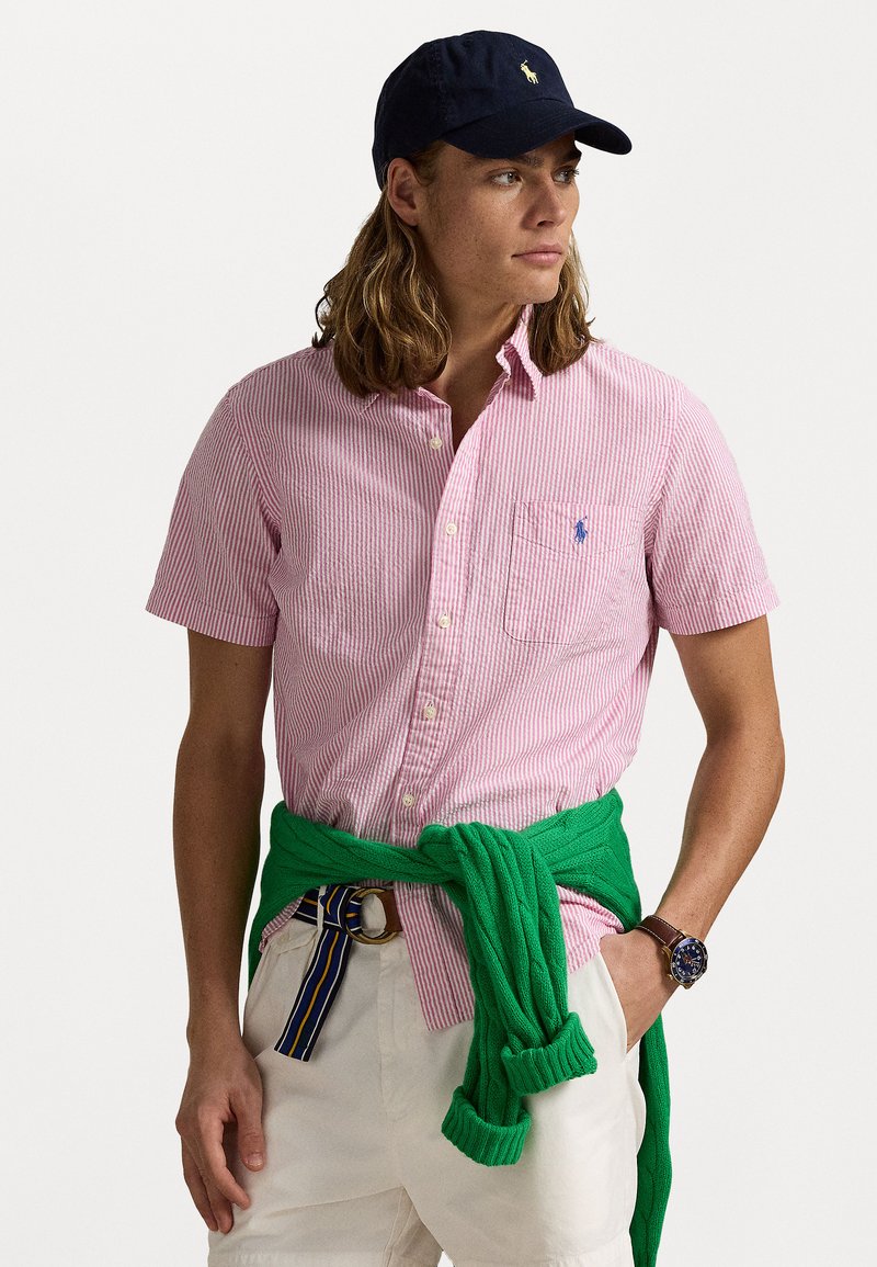 Červeno-bílé pruhované tričko s krátkým rukávem, modré logo na kapse, zelený svetr uvázaný v pase, béžové kalhoty, tmavá kšiltovka, viditelný náramkové hodinky.