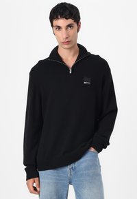 Maglione nero con zip, colletto e polsini a coste. Tessuto testurizzato e una piccola etichetta logo sul petto. Indossato con jeans azzurri chiari.