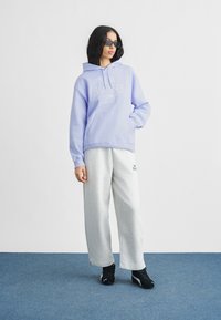 Ανοιχτό μοβ φούτερ με κουκούλα και λευκό κεντημένο λογότυπο, σε συνδυασμό με γκρι φόρμες. Το μοντέλο φοράει μαύρα αθλητικά παπούτσια και γυαλιά ηλίου μπροστά σε απλο φόντο.