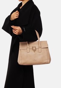Borsa a mano in pelle beige con finitura testurizzata, doppio manico e chiusura centrale. Abbinata a un abbigliamento esterno nero, presentata su uno sfondo bianco.