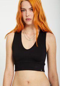 Top court noir à côtes avec un décolleté en V profond, présentant une coupe ajustée et un logo subtil sur le côté. Texture lisse.