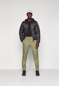 Black insulated jacket, black t-shirt, olive green joggers, and black athletic shoes; features a white visor with logo accents.