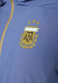 Μπλε αθλητικό μπουφάν με χρυσό έμβλημα της Ομοσπονδίας Ποδοσφαίρου της Αργεντινής, τέσσερα αστέρια και υφασμένους τόνους κατά μήκος των ραφών.