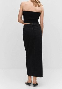 Falda maxi de mezclilla negra con corte recto y cinco bolsillos. Llevada con un top corto sin mangas negro y tacones abiertos negros. Textura suave.