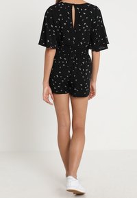 Combinaison noire à manches courtes, motif floral blanc, taille élastique, détail de dos ouvert et ourlet mi-thigh, associée à des baskets blanches.