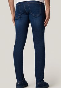 Mężczyzna w ciemnoniebieskich, dopasowanych dżinsach i granatowych sneakersach, stojący na jednolitym jasnym tle, z rękami swobodnie opartymi po bokach.