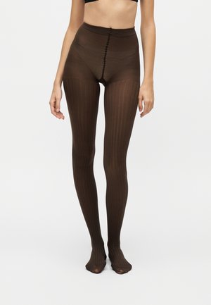 Femme portant des collants marron côtelés transparents, debout devant un fond uni clair, cadrée à la taille.
