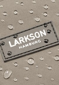 Vizes cseppek textúrált anyagon, közel egy fekete címkéhez, amelyen fehér "LARKSON HAMBURG" felirat található.