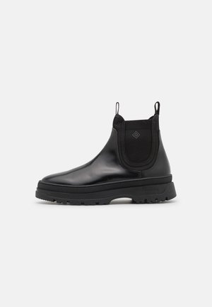 Bota Chelsea de cuero negro con suela gruesa de goma, paneles laterales elásticos y tiradores en el tobillo sobre fondo blanco.