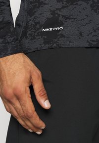 Schwarzes, langärmliges Sportoberteil mit einem dezenten strukturierten Muster und "NIKE PRO"-Logo, kombiniert mit eleganten schwarzen Hosen, wobei eine Hand an der Seite ruht.