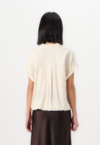 Light beige short-sleeve blouse with horizontal stripes, soft fabric, subtle pleat detail at the back, paired with a dark flowy skirt.