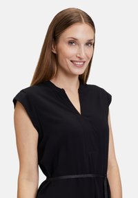 Schwarzes kurzärmeliges Kleid mit V-Ausschnitt, subtilen Knopfdetails und einer mit einem schmalen Band gebundenen Taillenakzent. Glattes, leichtes Material.