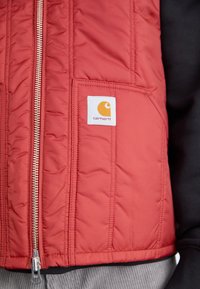 Rdeča oblazinjena brezrokavnik z zadrgo na sprednji strani, dvema žepoma in belim logotipom z oranžnim emblemom na levi strani prsnega dela.