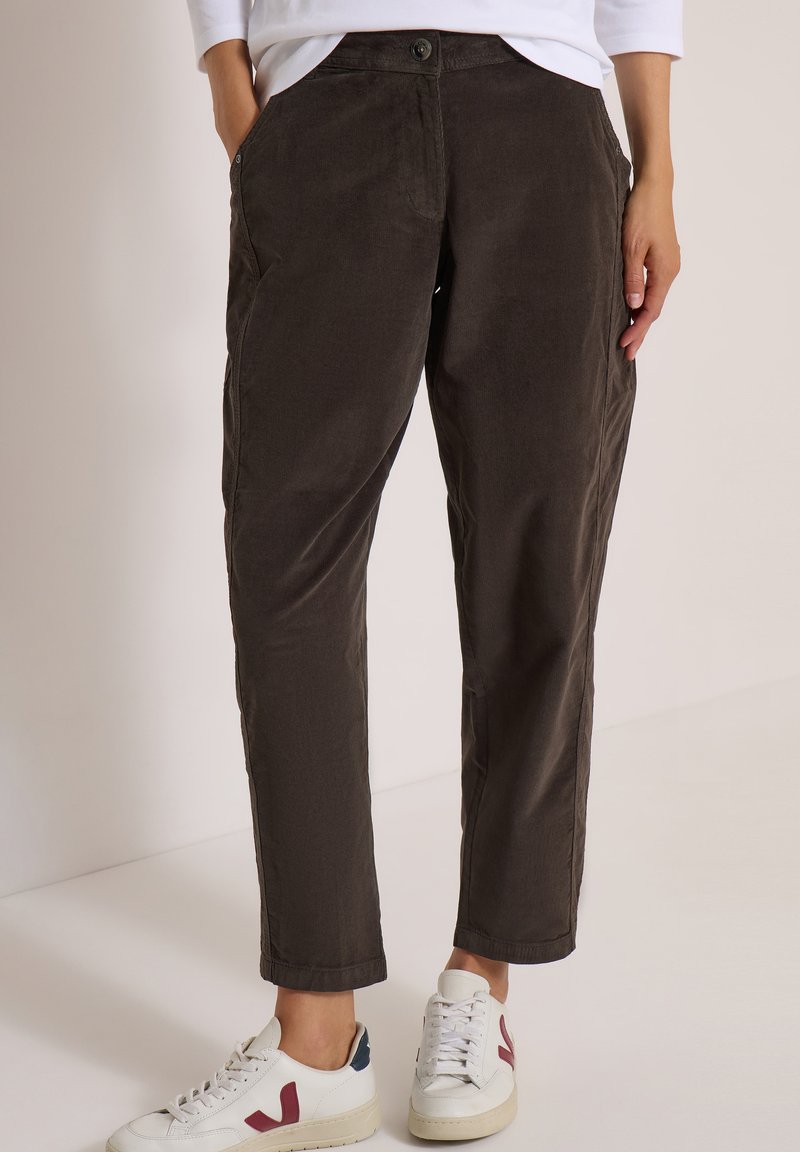 Femme portant un pantalon marron foncé fuselé et des baskets blanches avec des détails rouges et bleu marine, debout devant un fond clair uni.