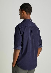 Camisa de botones de color azul marino con una textura suave, mangas remangadas y un collar. Presenta un pliegue en la parte trasera y un pequeño detalle de botón en el cuello.