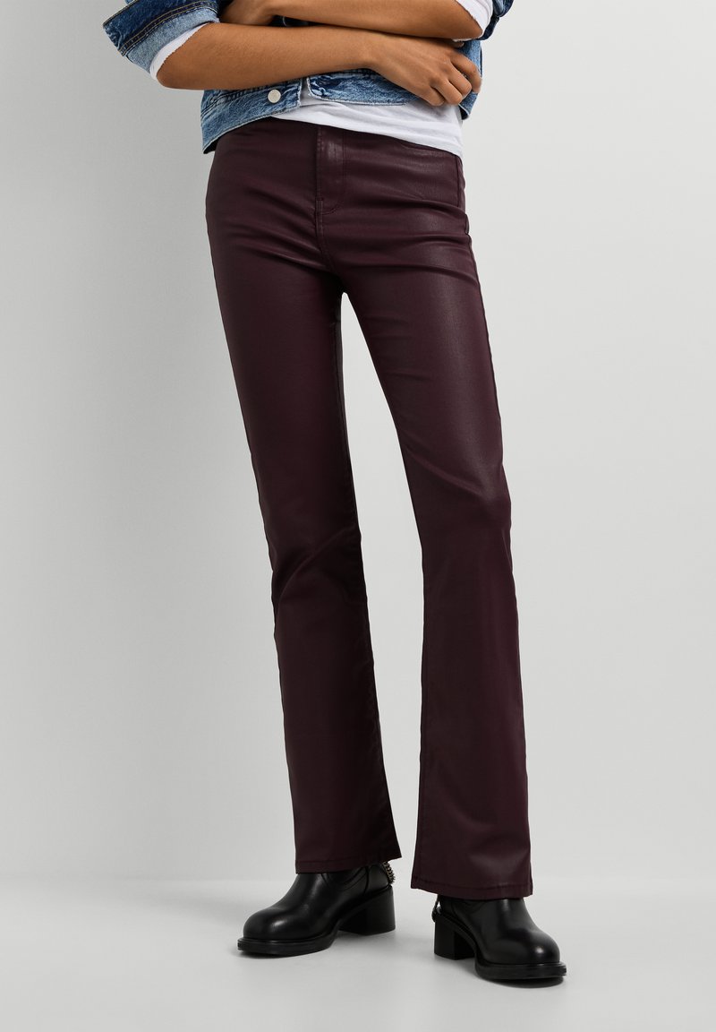 Person trägt dunkelviolette gerade Hose, schwarze Stiefel mit Absatz, weißes Hemd und Jeansjacke mit verschränkten Armen.
