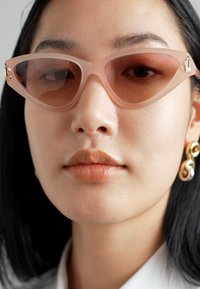 Cat-eye sunglasses in blush pink with translucent frames and brown tint. Gold hardware accent on the temple. Smooth texture.