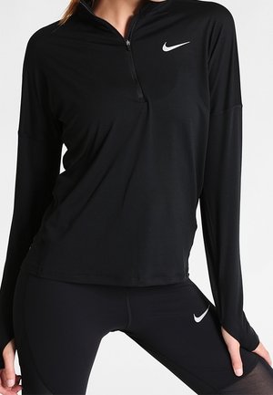 Kobieta ubrana w czarną bluzę Nike z długim rękawem i zamkiem oraz czarne legginsy, oba z białymi logo Nike, stoi z rękami na udach.