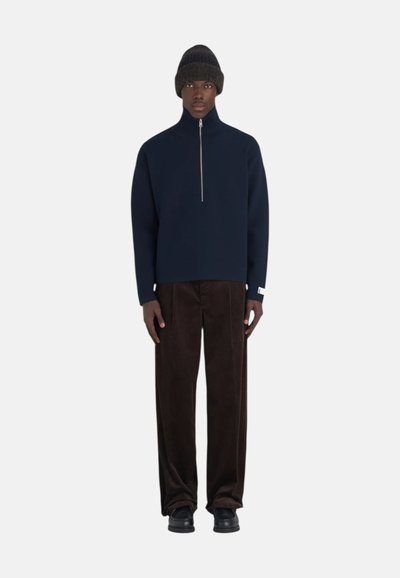 Pull-over bleu marine à col zippé, coupe décontractée, manches longues ; associé à un pantalon en velours côtelé marron et des chaussures noires ; le modèle porte un bonnet tricoté noir.