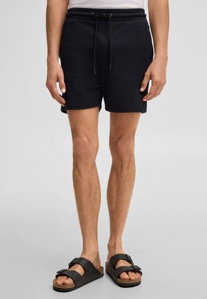 Mann mit schwarzen Shorts mit Kordelzug und schwarzen Sandalen mit Doppelriemen, stehend vor einem einfarbigen hellgrauen Hintergrund.