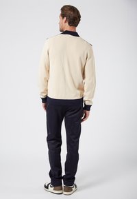 Beige ribbat stickad tröja med mörkblå krage och manschetter, tillsammans med mörkblå byxor och avslappnade sneakers, sedd bakifrån.