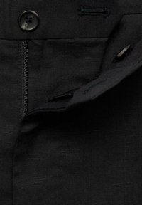 Tessuto nero con una superficie liscia, dotato di chiusura a pulsante, zip e un bordo ripiegato. Le cuciture sono ordinate e pulite.