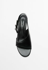 Zwarte leren slingback-sandaal met een brede, gebogen bovenste band en een cirkelvormige metalen accent. Gladde textuur en een open teen ontwerp.