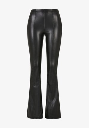 Rövid, göndör hajú nő fekete hosszú ujjú pólóban, fekete, magas derekú műbőr bőrnadrágban, és fekete, masszív bakancsban.