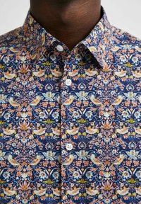 Košile na zapínání s tmavě modrým pozadím a květinovým a ptačím vzorem v pastelových barvách. Vyrobena z hladké látky s špičatým límcem.