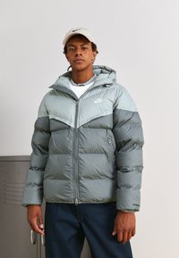 Casaco puffer cinza com capuz, apresentando um design bicolor e um logotipo no peito. Inclui fecho com zíper e bolsos laterais.