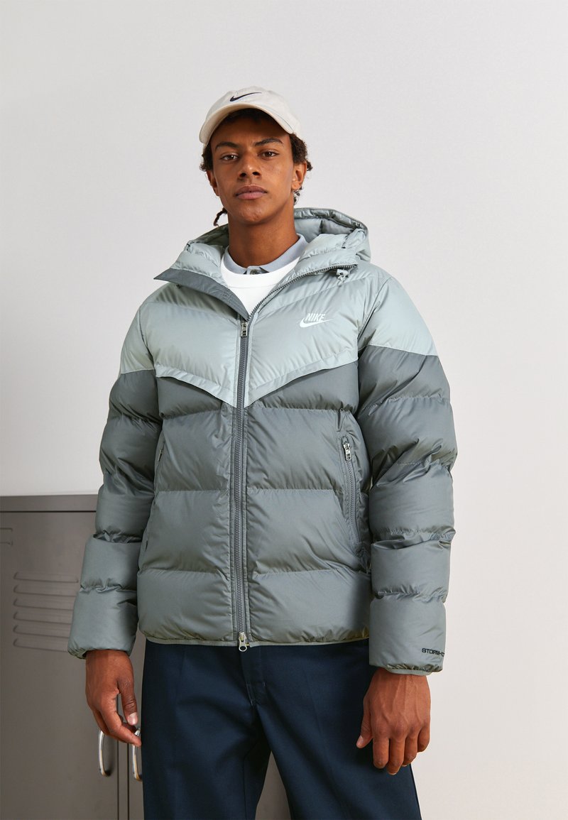 Casaco puffer cinza com capuz, apresentando um design bicolor e um logotipo no peito. Inclui fecho com zíper e bolsos laterais.