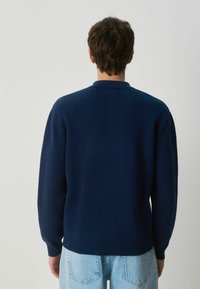 Marineblaues Strickpullover mit Polokragen, gerippten Bündchen und Saum. Die Rückansicht zeigt eine glatte Textur und eine lockere Passform. Denim-Jeans sind sichtbar.