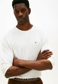 Uomo con le braccia incrociate che indossa una camicia bianca a maniche lunghe e pantaloni marroni con la cintura Tommy Hilfiger visibile, su sfondo bianco.