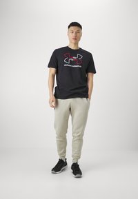 Sort bomulds t-shirt med rød og hvid Under Armour-logo, parret med lys grå sweatpants og sorte atletiske sko. Ensfarvet baggrund.