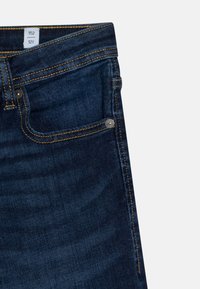 I jeans in denim blu scuro presentano cuciture gialle, una tasca anteriore con un rivetto argentato e una cintura etichettata con le taglie visibili.