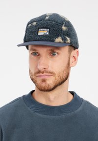 Cappello in pile con motivo blu e crema, dotato di una patch con logo frontale e visiera curva. Indossato con una felpa blu a girocollo.