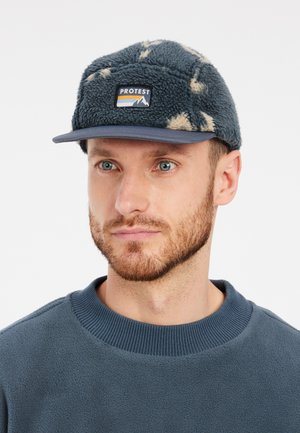 Fleece muts met een blauw en crèmekleurig patroon, voorzien van een logo patch aan de voorkant en een gebogen rand. Draagt bij een blauwe sweater met ronde hals.