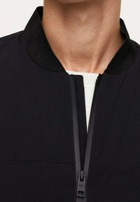 Veste noire avec col montant, fermeture éclair frontale et texture lisse ; comprend des détails de coutures horizontales.