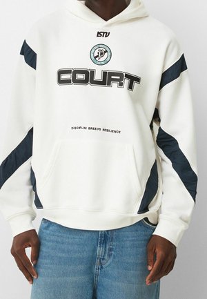 Hvid hættetrøje med sort tekst "COURT", cirkulært emblem, sorte diagonale striber på ærmerne og sætningen "DISCIPLINE BREEDS RESILIENCE", båret med blå jeans.