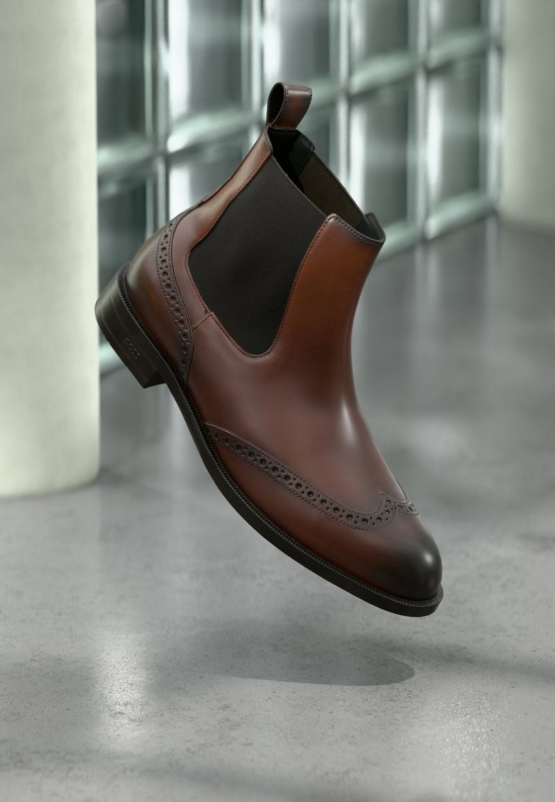 Botte Chelsea en cuir marron avec panneaux latéraux élastiques noirs et perforations décoratives, flottant au-dessus d'un sol en béton poli.